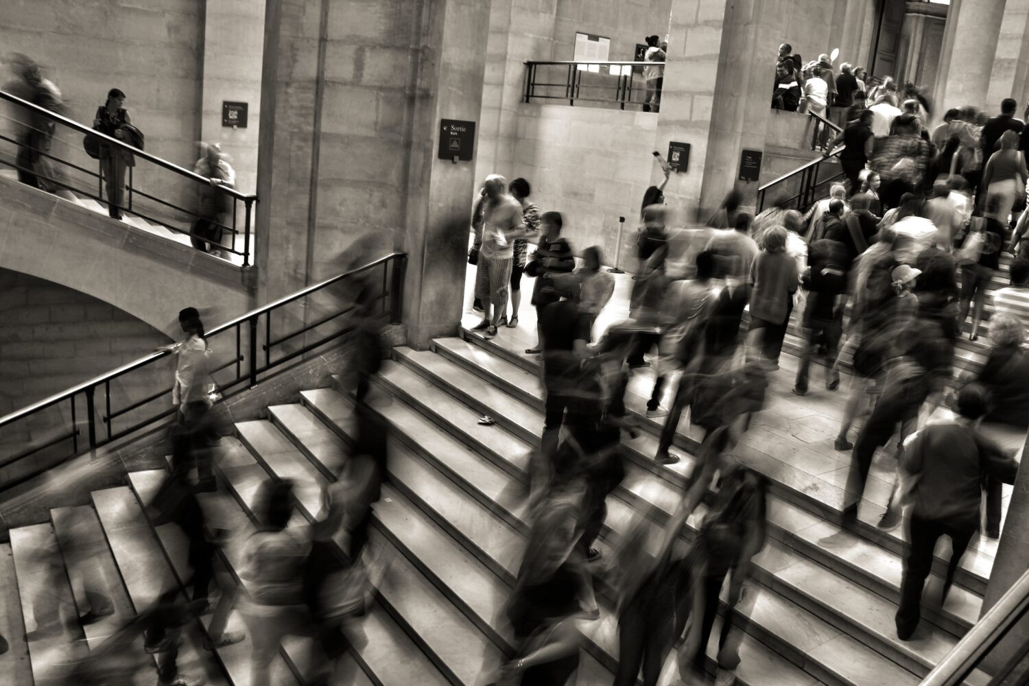 Greyscale photo of people in suits going up and down stairs, slightly blurred like they're moving too quickly for the photographer to capture