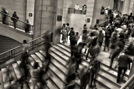 Greyscale photo of people in suits going up and down stairs, slightly blurred like they're moving too quickly for the photographer to capture